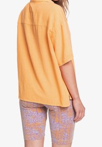 Camicia oversize a maniche corte in tessuto arancione brillante abbinata a pantaloni corti viola a fantasia con disegni gialli. Vestibilità casual e rilassata.