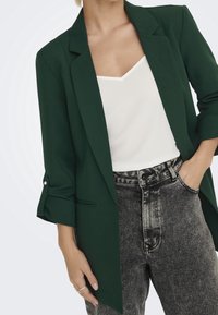 Grön kavaj med långa ärmar och knappar, bärs över en vit top och grå högmidjade jeans. Har en slät, strukturerad design.