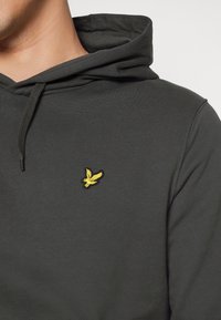 Sudadera gris oscura con tejido texturizado, bolsillo tipo canguro y un pequeño logotipo de un pájaro amarillo bordado en el lado izquierdo del pecho.