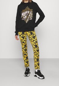 Felpa nera con un emblema decorativo dorato, abbinata a leggings neri e gialli con motivi barocchi. Sneakers nere completano l'outfit.