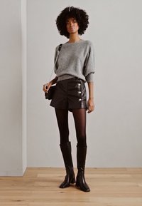 Pull en tricot gris, shorts en cuir noir avec des boutons sur le côté, collants noirs et bottes noires montantes. Le mannequin tient un petit sac à main noir.