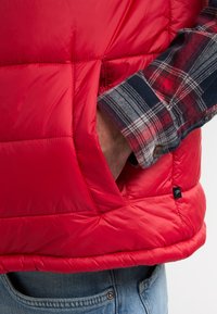 Rote gepolsterte Weste mit genähtem Design und einer Seitentasche. Der Träger hat einen Ärmel eines karierten Shirts sichtbar. Glatte, glänzende Stoffoberfläche.