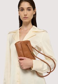 Brun læderhåndtaske med en glat tekstur, flad design og skulderrem; har en guldtonet hardwaredetalje.