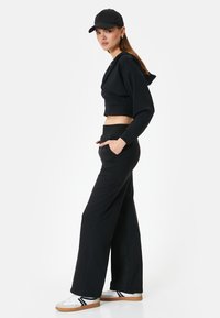 Schwarze, kurze Kapuzenjacke mit weiten schwarzen Hosen und weißen Sportschuhen. Verfügt über einen elastischen Bund und Seitentaschen.