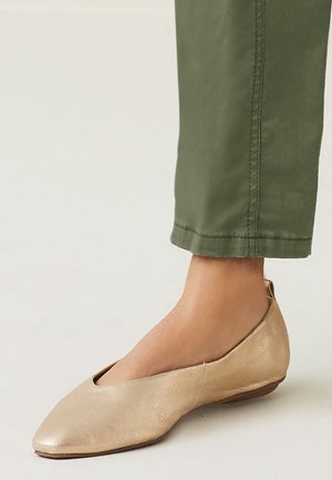 Metallisch goldene Ballerinas mit spitzem Zehenbereich, niedrigem Absatz und Ausschnitten am oberen Teil. Getragen mit olivgrünen, cropped Hosen.