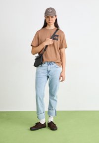 Braunes Baumwoll-T-Shirt mit schwarzem Logo, hellblaue Jeans mit umgeschlagenem Saum an den Knöcheln, schwarze Umhängetasche, graue Mütze und braune Schuhe mit weißen Socken.