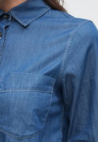 Gros plan d'une chemise en denim bleue avec poche avant, boutons noirs et col pointu sur une personne aux cheveux foncés.