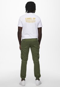 Vit t-shirt med "CAMEL UP" tryckt i gult på baksidan, tillsammans med olivgröna cargobyxor och vita sneakers.