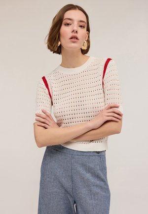 Maglione lavorato a maglia bianco con accenti rossi, caratterizzato da un design perforato, maniche corte e scollatura rotonda, abbinato a pantaloni azzurri chiari.