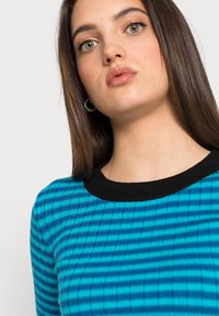 Pull en tricot rayé bleu et teal avec un col côtelé noir, présentant une coupe ajustée et des lignes verticales texturées.