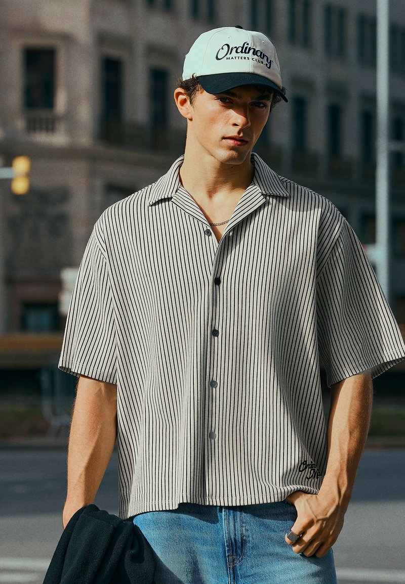 Jeune homme portant une casquette blanche et noire, une chemise à manches courtes rayée et un jean bleu, tenant une veste sombre dans un environnement urbain.