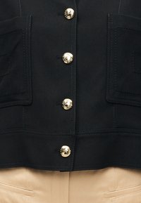 Κοντινή λήψη μαύρου σακακιού με πέντε χρυσά κουμπιά και δύο εμπρόσθιες τσέπες, φορεμένο πάνω από μπεζ παντελόνια.