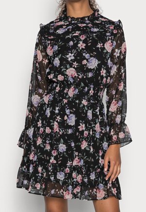 Czarna sukienka w kwiaty z przezroczystymi długimi rękawami, marszczonymi mankietami, elastyczną talią i falbaną u dołu, noszona przez stojącą osobę.