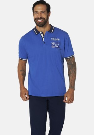 Mann lächelt, trägt ein blaues Poloshirt mit dem Text "Nordic Sunrise" und dunkle Hosen, zeigt Tattoos auf beiden Unterarmen, steht vor einem einfarbigen Hintergrund.