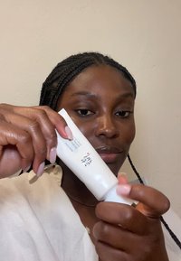 Vit kosmetisk tub som hålls i handen, cylindrisk form med svart och röd text på etiketten, slät yta, matt finish. Neutral bakgrund.