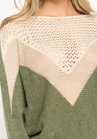 Vrouw in een gebreide trui met een groene basis, beige chevronpatroon en een crèmekleurige opengehaakte yoke, die haar bovenlichaam en blonde haar toont.