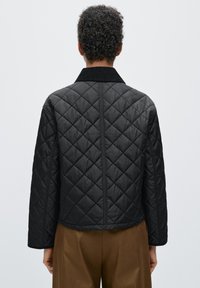 Zwarte doorgestikte jas met ruitpatroon, met een zwarte kraag en lange mouwen, gedragen bij een bruine broek, gezien vanaf de achterkant.