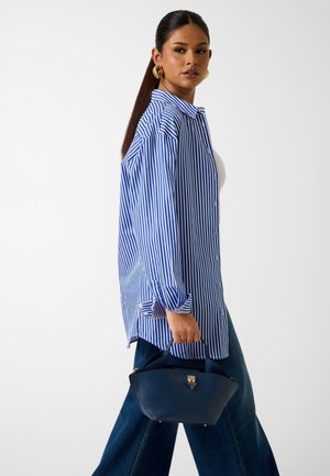Woman in blue and white vertical striped oversized shirt, dark wide-leg jeans, holding a navy blue handbag with gold clasp.
