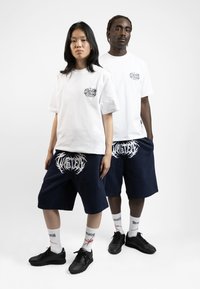 T-shirts blancs à manches courtes avec un graphique noir sur la poitrine, associés à des shorts bleu marine portant un texte logo blanc, des baskets noires et des chaussettes à motifs.