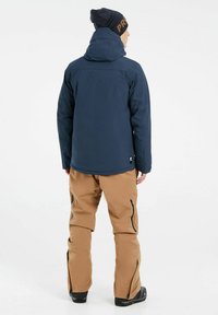 Chaqueta impermeable de color azul marino con capucha, combinada con pantalones aislantes color beige que cuentan con bolsillos con cremallera y cremalleras en los tobillos, y llevada con botas negras.