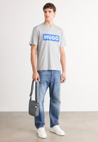 Pilkas trumpas rankovėmis marškinėliai su mėlynu "HUGO" logotipu, derinami su šviesiai mėlynomis džinsais ir baltais sportbačiais. Laikoma maža pilka petinė rankinė.