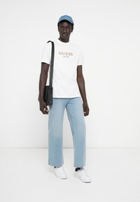 T-shirt bianca a maniche corte con grafica arancione "GUESS EST. 1981", jeans dritti blu chiaro, borsa a tracolla nera e sneakers bianche.