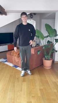 Ung mann som smiler, står i stuen med tregulv, stor potteplante, oransje sofa og TV i bakgrunnen.