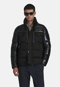 Zwarte pufferjack met glanzende schouders en een matte body, is voorzien van dubbele borstzakken, drukknoopsluitingen en een hoge kraag. Draag het met een zonnebril.