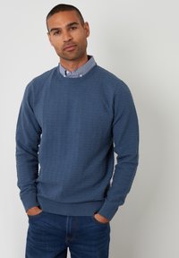 Blauwe textuurtrui met een ronde halslijn, geribbelde manchetten en zoom, gecombineerd met een overhemd met kraag eronder, gedragen met denim jeans.