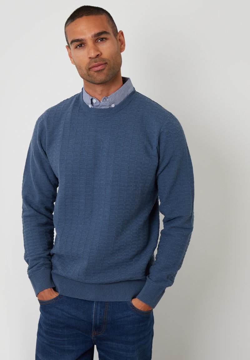 Blauwe textuurtrui met een ronde halslijn, geribbelde manchetten en zoom, gecombineerd met een overhemd met kraag eronder, gedragen met denim jeans.