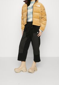 Person som bär en tan puffjacka, blå mönstrad topp, svarta byxor med lappar och beige klumpiga plattformsskor, stående på en ljusgrå golv.