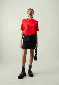T-shirt vermelho de manga curta com o texto "FEMME", combinado com uma saia de ganga preta e botins de plataforma pretos. A modelo segura uma pequena bolsa preta.