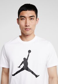 Hvid bomulds t-shirt med en stor sort silhuet af en basketballspiller i et hoppe-pose på forsiden. Korte ærmer og rund halsudskæring.