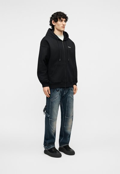 Νεαρός με σγουρά μαλλιά, που φορεί μαύρο φούτερ με φερμουάρ, λευκό μπλουζάκι, ξεβαμμένα μπλε cargo τζιν και μαύρα αθλητικά παπούτσια, στέκεται μπροστά σε μια απλή φόντο.