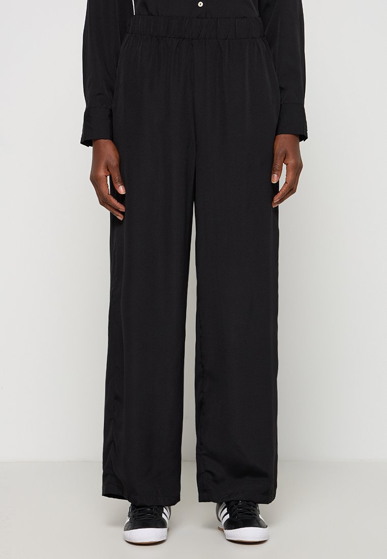 Zwarte broek met wijde pijpen en een elastische tailleband, gemaakt van een gladde stof, gecombineerd met zwarte sneakers met witte strepen.