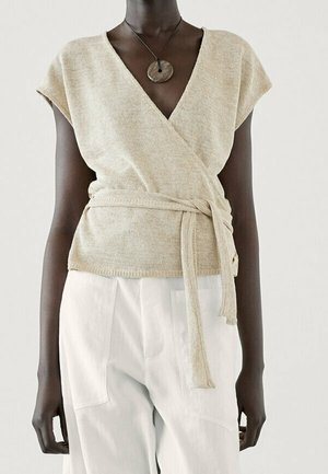 Dark-skinned person wearing a beige wrap knit top with short sleeves and a tie belt, paired with loose white pants, and a round stone necklace.