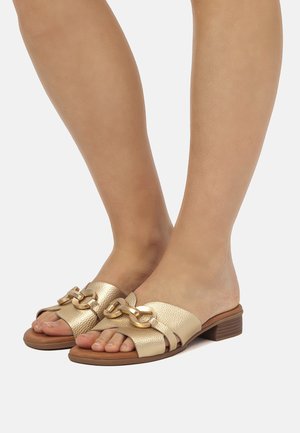 Piedi con sandali aperti, slip-on dorati con texture e dettaglio di catena chunky, tacco basso impilato in legno su uno sfondo semplice.