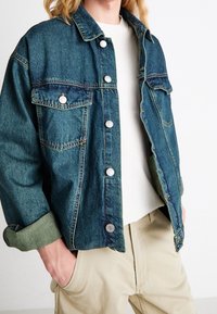 Spijkerjack in donkerblauw met zilverkleurige drukknopen, borstzakken en opgerolde manchetten, gecombineerd met een wit shirt en beige broek.