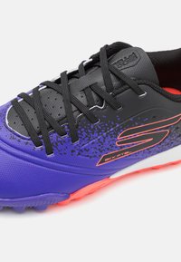 Chaussure de football violette avec des accents noirs et une tige texturée. Présente des lacets noirs, une semelle rose et une semelle intermédiaire blanche.
