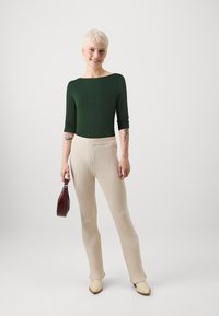 Green fitted top, cream ribbed trousers, yellow heels, holding a textured burgundy handbag. Simple lines, smooth fabrics, and neutral colors.