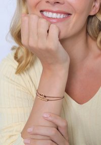 Bracciale in corda marrone con chiusura circolare in oro e accento testurizzato in oro, indossato su un polso su uno sfondo chiaro.