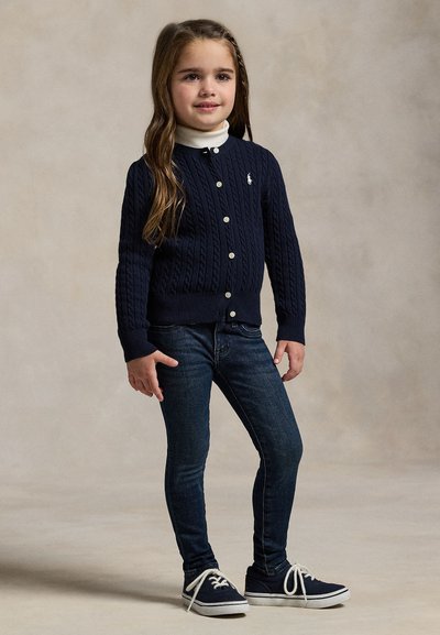 Ragazza giovane in piedi che indossa un cardigan blu navy, dolcevita bianco, jeans scuri e scarpe da ginnastica blu navy con lacci bianchi su uno sfondo semplice.