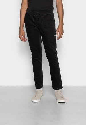 Hombre con pantalones casuales negros con cordón en la cintura y zapatillas beige, de pie contra un fondo claro liso.