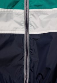 Färgad jacka med blockering som har paneler i teal, vitt och marinblått, med en central dragkedja och kontrasterande sömmar. Tillverkad av ett slätt, lätt tyg.