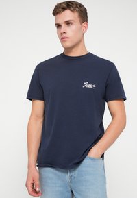 Joven con cabello corto vestido con una camiseta de Guess azul marino y jeans celestes, de pie con una mano en el bolsillo frente a un fondo blanco.