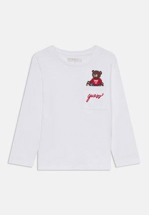 Biała, długorękawowa koszulka z organicznej bawełny, z haftowanym niedźwiedziem na kieszeni i czerwonym logo "guess" poniżej.