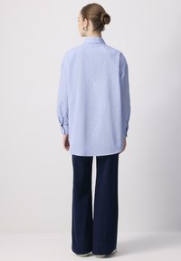 Donna con i capelli raccolti in un bun, che indossa orecchini a cerchio in argento, una camicia a righe verticali blu e bianca e pantaloni scuri a gamba larga, voltata di spalle.