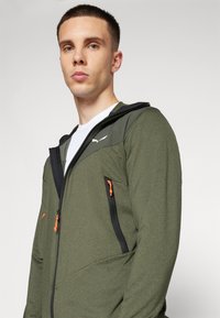 Πράσινο μπουφάν με φερμουάρ και μαύρες λεπτομέρειες, που διαθέτει πορτοκαλί λαβές φερμουάρ και υφή υφάσματος. Σχεδίαση με κουκούλα και τσέπες.