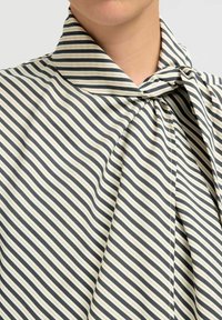 Nærbillede af en person iført en sort, hvid og beige vertikalt stribet bluse med en knyttet høj krave.