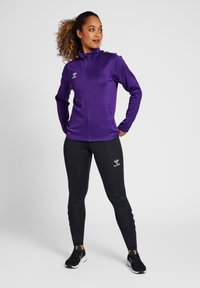 Lilla treningsjakke med glidelås foran og høy krage, båret over svarte leggings. Logomerking på begge plaggene, glatt stofftekstur.